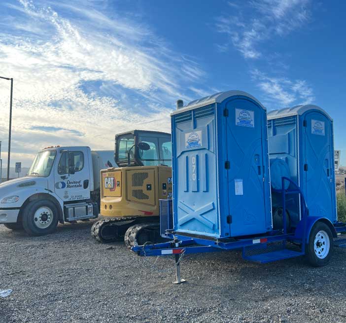 A blue double towable restroom unit parked alongside construction equipment on a job site to provide mobile site services, following osha compliance for job sites