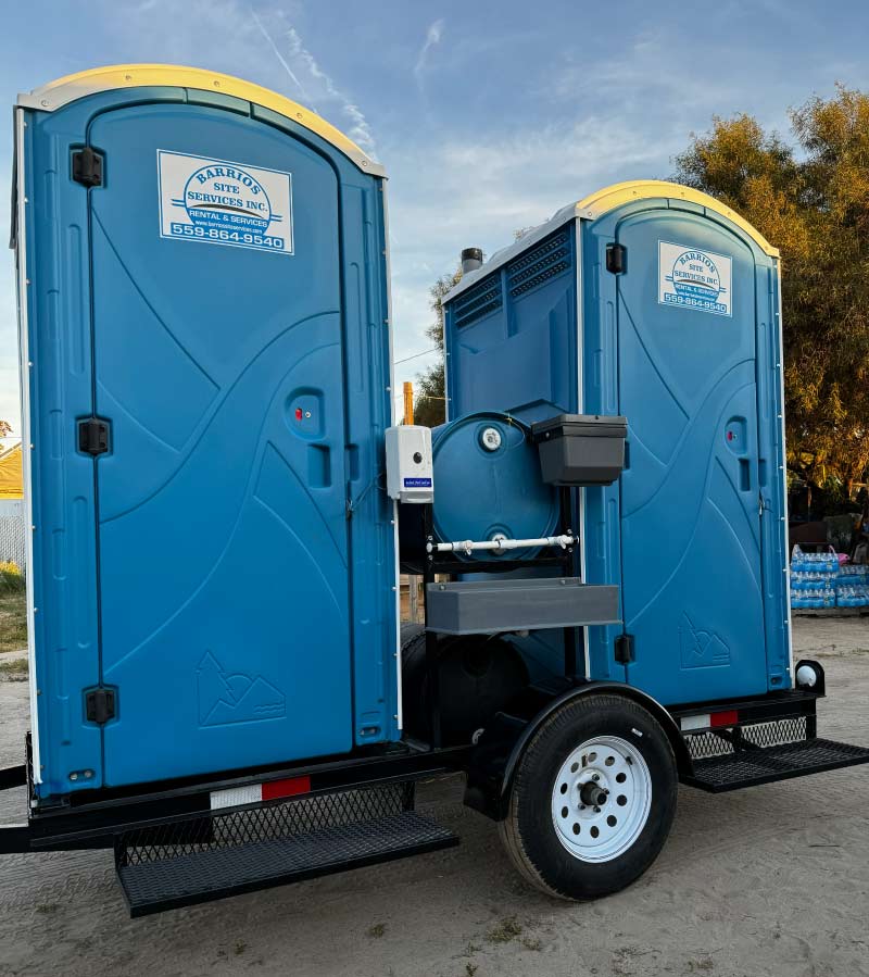 A pair of blue Barrios Site Services towable portable restrooms on a heavy-duty trailer, equipped with specialized handwashing stations for remote job sites and agricultural field workers.
