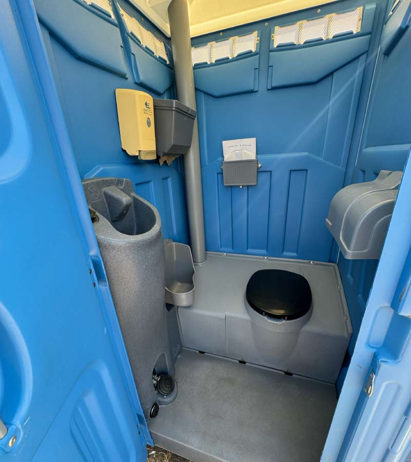 Interior view of a clean Barrios Site Services deluxe portable toilet featuring an integrated foot-pump operated handwash station, soap dispenser, paper towel holder, and a 3-roll toilet paper dispenser.