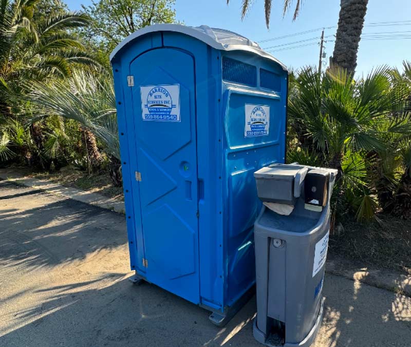 A high-visibility blue single-stall portable toilet and a standalone grey handwash station positioned on a paved pathway surrounded by tropical palm trees.