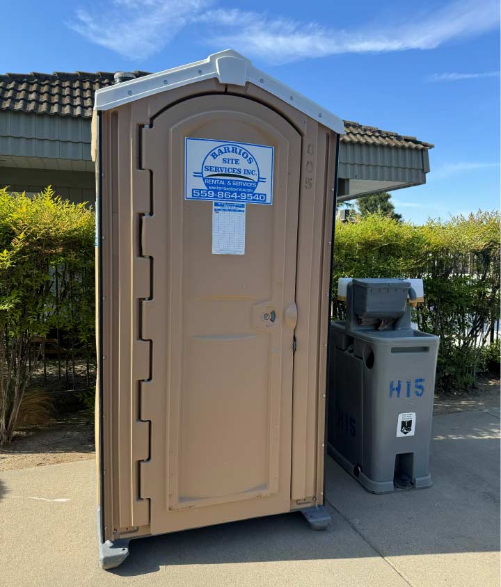 A deluxe tan portable restroom featuring an up-to-date service log, placed adjacent to a premium grey handwashing station on a clean concrete surface.