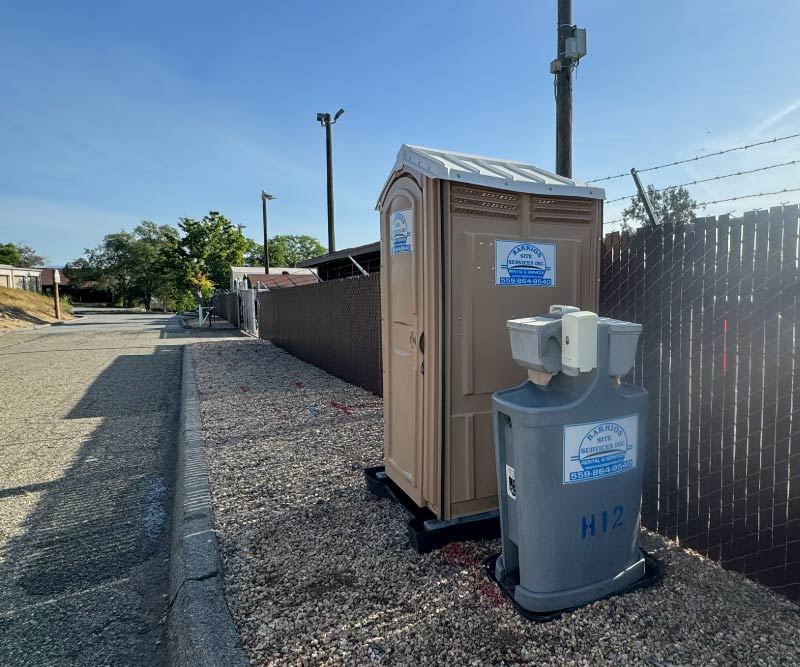 A clean, tan Barrios Site Services portable toilet paired with a standalone grey dual-basin handwashing station, professionally positioned on a gravel shoulder next to a secure perimeter fence.