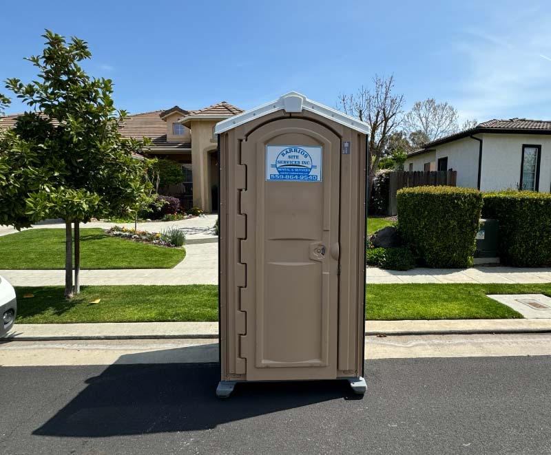 A pristine, beige event-ready portable toilet placed curbside on a clean asphalt street in front of a modern suburban home and manicured green lawn, for Porta Potty Rentals for Film & Production