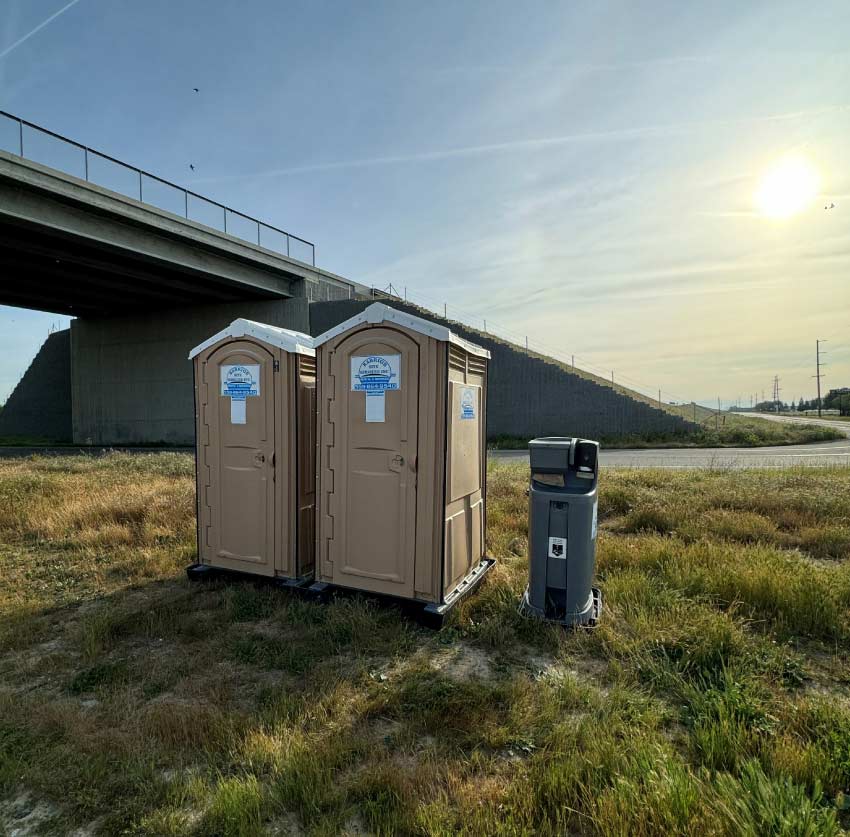 A deluxe tan portable restroom featuring an up-to-date service log, placed adjacent to a premium grey handwashing station on a clean concrete surface.