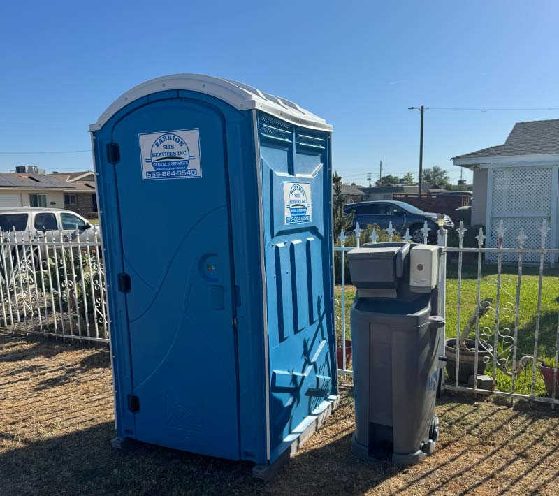A vibrant blue portable restroom unit from Barrios Site Services standing alongside a grey outdoor hand sanitizing station in a well-maintained residential neighborhood.
