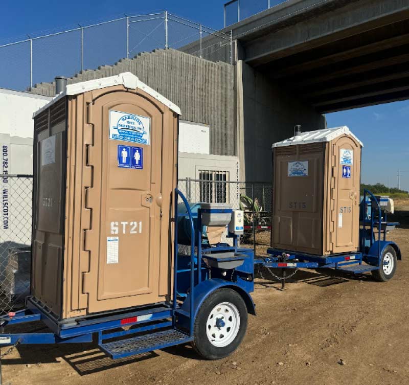 Two mobile, towable tan portable restrooms securely mounted on heavy-duty blue single-axle trailers, staged for rapid deployment beneath a concrete highway overpass, for porta potty rentals for sports events